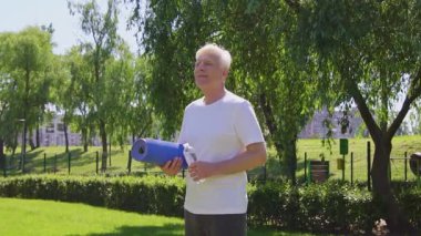 Beyaz tişörtlü atletik yaşlı adam yazın parkta yoga minderi ve su şişesi tutuyor, aktif gri saçlı emekli dışarıda egzersiz yaparken gülümsüyor.