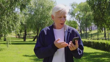 Yaşlılar akıllı telefon kullanır ve parkta şaşırır. Yaşlı, gri saçlı emekli cep telefonuna hayret içinde bakar.