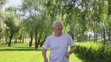 Beyaz tişörtlü yaşlı sporcu yazın parkta koşuyor ve gülümsüyor, aktif gri saçlı emekli spor yapıyor ve doğada aktif olarak rahatlıyor.