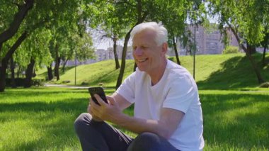 Yaşlı beyefendi parkta oturuyor ve akıllı telefon kullanıyor. Yaşlı, gri saçlı emekli cep telefonuna bakıyor ve doğada gülümsüyor.