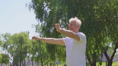 Beyaz tişörtlü atletik yaşlı adam yazın parkta egzersiz yapıyor ve gülümsüyor, aktif gri saçlı emekli spor yapıyor ve eklemlerini esnetiyor.