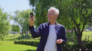 Yaşlı adam akıllı telefondan konuşuyor ve parkta gülüyor. Neşeli, gri saçlı emekli dışarıda video aracılığıyla iletişim kuruyor.