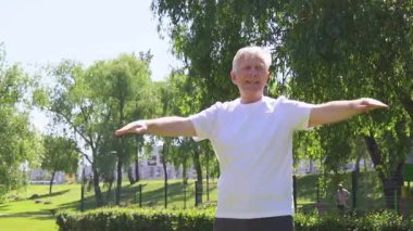 Beyaz tişörtlü atletik yaşlı adam yazın parkta egzersiz yapıyor ve gülümsüyor, aktif gri saçlı emekli spor yapıyor ve doğada aktif olarak rahatlıyor.