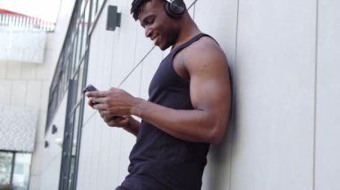 Afro-Amerikalı adam akıllı telefon kullanıyor ve sokakta müzik dinliyor, atletik adam cep telefonuyla yazı yazıyor ve şehir geçmişine gülümsüyor.