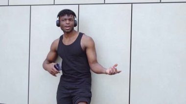 Afro-Amerikan atletik adam akıllı telefon kullanıyor ve sokakta müzik dinliyor, kaslı adam şehir arka planında gülümsüyor.