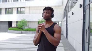 Spor giyim sektöründe Afrikalı Amerikalı bir adam esniyor ve sokakta müzik dinliyor, atletik adam ortak egzersizler yapıyor ve kulaklıkla egzersiz yapıyor.