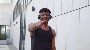 Kulaklıklı Afro-Amerikalı adam kamerayı işaret ederken alay ediyor ve gülüyor. Sportif adam sizi şehir sokaklarında suçluyor ve alay ediyor.