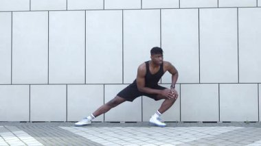 Spor giyim kuşağında Afrikalı Amerikalı bir adam bacaklarını şehir duvarına dayayıp egzersiz yapıyor, atletik bir adam dışarıda egzersiz yapıyor.