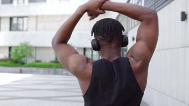 Spor giyim sektöründe Afrikalı Amerikalı bir adam esniyor ve sokakta müzik dinliyor, atletik adam ortak egzersizler yapıyor ve kulaklıkla egzersiz yapıyor.