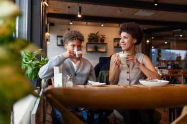 Afro-Amerikalı kadın ve oğlu kafede oturup kahve içiyorlar ve gülümsüyorlar, kıvırcık saçlı genç ve annesi restoranda öğle yemeği yiyorlar.