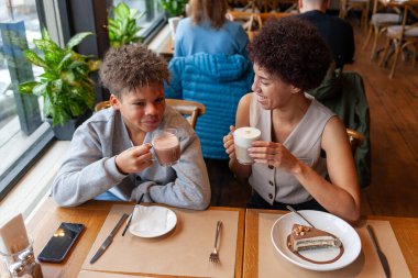 Afro-Amerikalı kadın ve oğlu kafede oturup kahve içiyorlar ve gülümsüyorlar, kıvırcık saçlı genç ve annesi restoranda öğle yemeği yiyorlar.