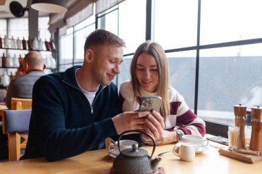Genç çift kafedeki masada oturuyor ve akıllı telefon kullanıyor, kız ve erkek restoranda çay içiyor, cep telefonlarına bakıyor ve gülümsüyor.