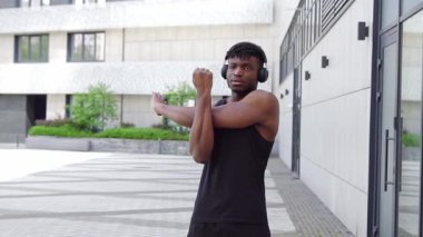 Spor giyim sektöründe Afrikalı Amerikalı bir adam esniyor ve sokakta müzik dinliyor, atletik adam ortak egzersizler yapıyor ve kulaklıkla egzersiz yapıyor.