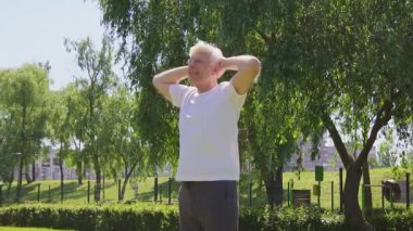 Beyaz tişörtlü atletik yaşlı adam yazın parkta egzersiz yapıyor ve gülümsüyor, aktif gri saçlı emekli spor yapıyor ve doğada aktif olarak rahatlıyor.