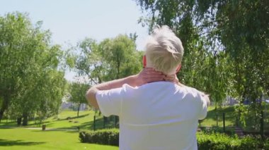 Beyaz tişörtlü atletik yaşlı adam yazın parkta egzersiz yapıyor ve gülümsüyor. Aktif gri saçlı emekli, spor yapıyor ve boynuna masaj yapıyor.