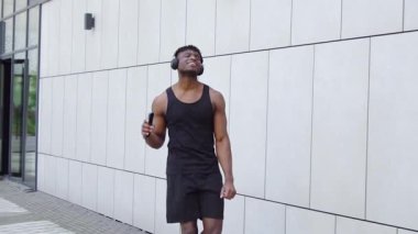 Sokakta müzik dinleyen ve dans eden Afrikalı Amerikalı atletik adam, cep telefonlu kaslı adam şehir arka planında başarıyı kutluyor ve seviniyor.