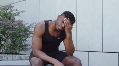 Düşünceli Afro-Amerikalı bir adam spor kıyafetleriyle sokakta oturuyor ve üzgün hissediyor, sayısız sporcu bir karar vermeyi ve acı çekmeyi düşünüyor.