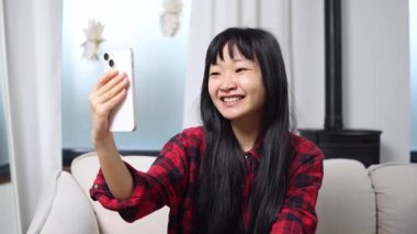 Asyalı genç bir kadın evde telefonla konuşuyor ve selamlıyor, Koreli bir kız kanepede görüntülü konuşuyor ve gülümsüyor.