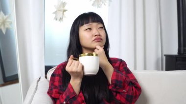 Asyalı düşünceli kadın kanepede oturup çay içerken düşünüyor, elinde fincanla hayal kuruyor ve evde hayal kuruyor.