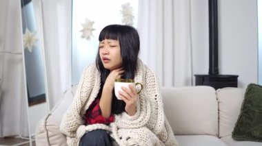 Battaniyeyle kaplanmış Asyalı genç bir kadın grip ve ateşten muzdarip, hasta kız evde çay içiyor ve soğuk davranıyor.