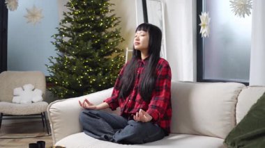 Genç Asyalı kadın evdeki kanepede meditasyon yaparken ve nefes alma alıştırması yaparken Koreli kız lotus pozisyonunda oturup yoga yapıyor.