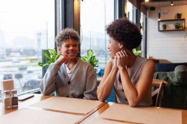 Afro-Amerikalı bir kadın ve oğlu kafede oturmuş kıvırcık saçlı bir ergenle konuşuyorlar ve annesi restoranda oturmuş gülümsüyor.