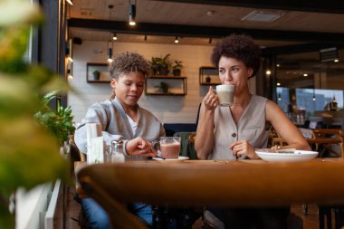 Afro-Amerikalı kadın ve oğlu kafede oturup kahve içiyorlar ve gülümsüyorlar, kıvırcık saçlı genç ve annesi restoranda öğle yemeği yiyorlar.