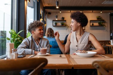 Afro-Amerikalı kadın ve oğlu kafede oturup kahve içip tatlı yiyorlar. Kıvırcık saçlı genç ve annesi restoranda öğle yemeği yiyorlar.