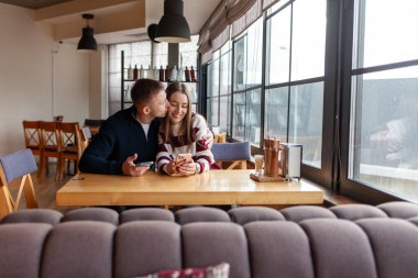 Genç çift kafedeki masada oturuyor ve akıllı telefon kullanıyor, kız ve erkek restoranda çay içiyor, cep telefonlarına bakıyor ve gülümsüyor.