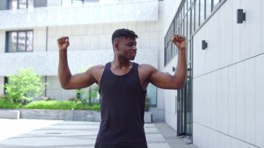Spor giyim sektöründe Afro-Amerikalı bir adam bisepslerini gösteriyor ve kameraya gülümsüyor, atletik adam güç gösterisi ve kentsel geçmiş üzerine eğitim gösteriyor.