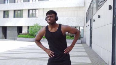 Spor giyim sektöründe Afrikalı Amerikalı bir adam esniyor ve sokakta müzik dinliyor, atletik adam ortak egzersizler yapıyor ve kulaklıkla egzersiz yapıyor.