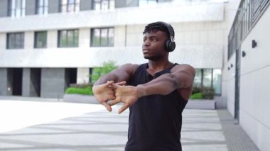 Spor giyim sektöründe Afrikalı Amerikalı bir adam esniyor ve sokakta müzik dinliyor, atletik adam ortak egzersizler yapıyor ve kulaklıkla egzersiz yapıyor.