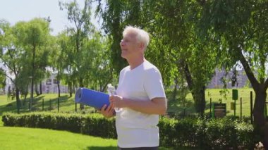 Beyaz tişörtlü atletik yaşlı adam yazın parkta yoga minderi ve su şişesi tutuyor, aktif gri saçlı emekli dışarıda egzersiz yaparken gülümsüyor.