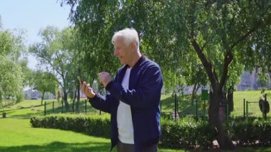 Yaşlı adam akıllı telefon kullanıyor ve parkta online olarak kazanıyor. Gri saçlı emekliler açık havada zaferini kutluyor.