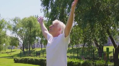 Beyaz tişörtlü yaşlı atletik adam yazın parkta meditasyon yapıyor. Aktif gri saçlı emekliler doğada nefes alıyor.
