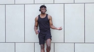 Afro-Amerikan atletik adam akıllı telefon kullanıyor ve sokakta müzik dinliyor, kaslı adam şehir arka planında gülümsüyor.