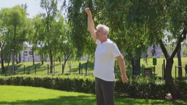 Beyaz tişörtlü atletik yaşlı adam yazın parkta egzersiz yapıyor ve gülümsüyor, aktif gri saçlı emekli spor yapıyor ve doğada aktif olarak rahatlıyor.