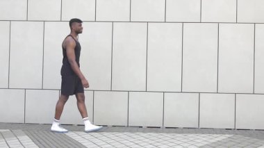 Spor kıyafetli Afrikalı bir adam duvarın arkasında yürüyor, atletik bir adam şehirde yürüyor ve gülümsüyor.