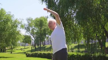 Beyaz tişörtlü atletik yaşlı adam yazın parkta egzersiz yapıyor ve gülümsüyor, aktif gri saçlı emekli spor yapıyor ve doğada aktif olarak rahatlıyor.