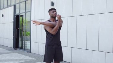 Spor giyim sektöründe Afrikalı Amerikalı bir adam esniyor ve sokakta müzik dinliyor, atletik adam ortak egzersizler yapıyor ve kulaklıkla egzersiz yapıyor.