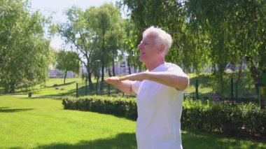 Beyaz tişörtlü atletik yaşlı adam yazın parkta egzersiz yapıyor ve gülümsüyor, aktif gri saçlı emekli spor yapıyor ve doğada aktif olarak rahatlıyor.