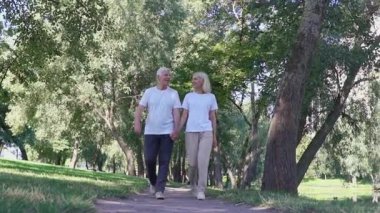Parkta el ele tutuşan yaşlı çift, gri saçlı adam ve kadın ormanda yürüyor ve konuşuyor ve gülümsüyor.
