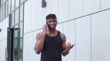 Afro-Amerikalı sporcu telefonda konuşuyor ve sokakta gülümsüyor, atletik adam cep telefonuyla konuşuyor şehir planına karşı.