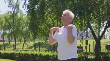 Beyaz tişörtlü atletik yaşlı adam yazın parkta egzersiz yapıyor ve gülümsüyor, aktif gri saçlı emekli spor yapıyor ve doğada aktif olarak rahatlıyor.