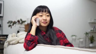 Genç Asyalı kadın kanepede oturuyor ve telefonla konuşuyor, cep telefonuyla kız iletişimi kuruyor ve evde gülümsüyor.