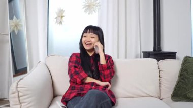Genç Asyalı kadın kanepede oturuyor ve telefonla konuşuyor, cep telefonuyla kız iletişimi kuruyor ve evde gülümsüyor.