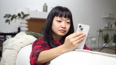 Asyalı genç bir kadın kanepede oturmuş gülümsüyor ve akıllı telefon kullanıyor. Kız telefona bakıyor ve evde gülüyor.