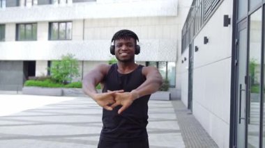 Spor giyim sektöründe Afrikalı Amerikalı bir adam esniyor ve sokakta müzik dinliyor, atletik adam ortak egzersizler yapıyor ve kulaklıkla egzersiz yapıyor.