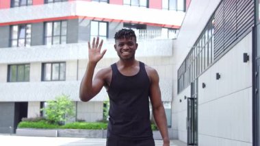 Afro-Amerikan spor giyim kuşağı giymiş, caddede hoş geldin jesti yapan atletik bir adam selam veriyor, el sallıyor ve şehir geçmişinde sarılıyor.
