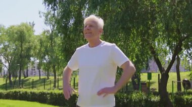 Beyaz tişörtlü sportif yaşlı adam yazın kollarını kavuşturup parkta duruyor ve gülümsüyor. Doğada aktif gri saçlı bir emeklinin portresi.